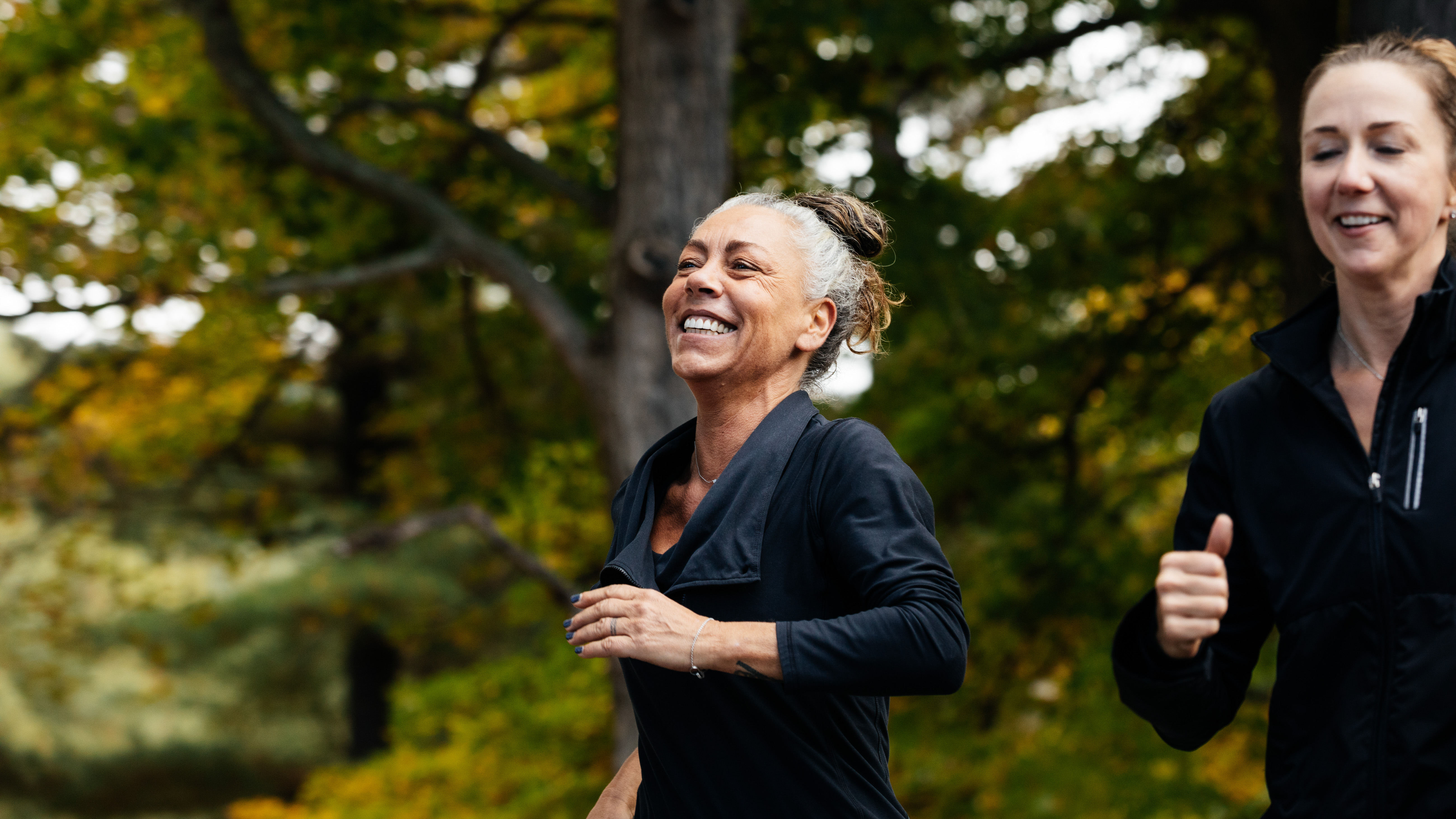 Two women run through a leafy park wearing hooded sports tops and smiling.