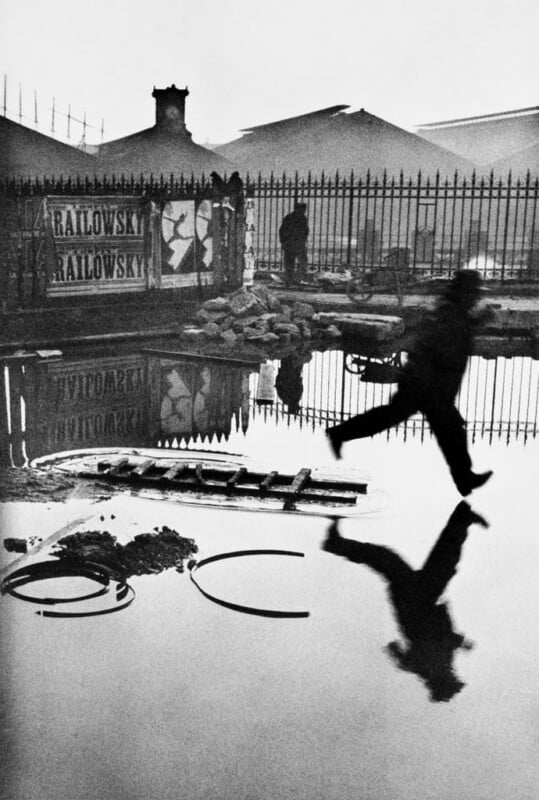 A person jumps over a large puddle next to a fence, their reflection visible in the water. In the background, a person stands near a poster-covered wall and there are industrial buildings and scattered debris. The scene is black and white.