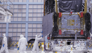 People wearing white coveralls and masks work around a giant covered piece of machinery in a cleanroom warehouse