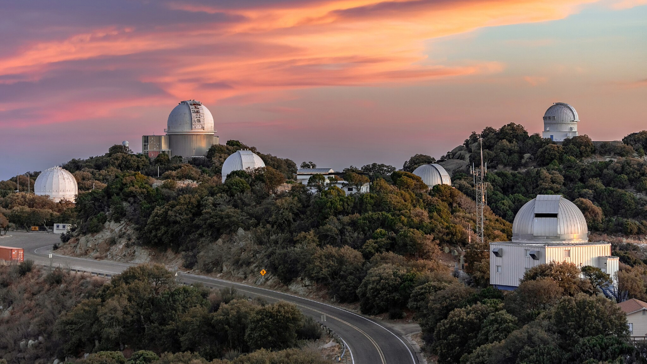 A series of domed buildings sit among rocks and trees with a bright pink sunset behind them
