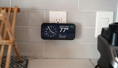 A smartphone mounted horizontally on a kitchen wall outlet displaying a clock and weather information showing 77°F and mostly sunny in Atlantic Highlands. The background includes white tiled walls, a wooden dish rack on the left, and a black kitchen appliance on the right.