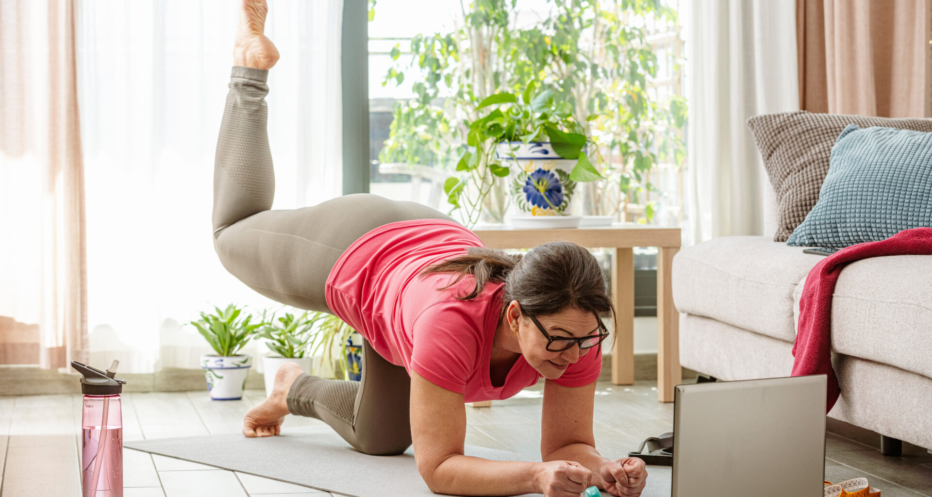 A woman practices Pilates at home in her living room on a mat. She is balancing, close to the ground, on her forearms and her bent left knee. Her right leg is bent at 90° and elevated behind her. In the room we see a wtaer bottle, a couch, a plant and a coffee table. A laptop is open in front of her.