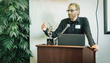 A speaker stands at a podium giving remarks during a Cal State LA Biostart event, with an award trophy and laptop on the podium and a potted plant in the background.