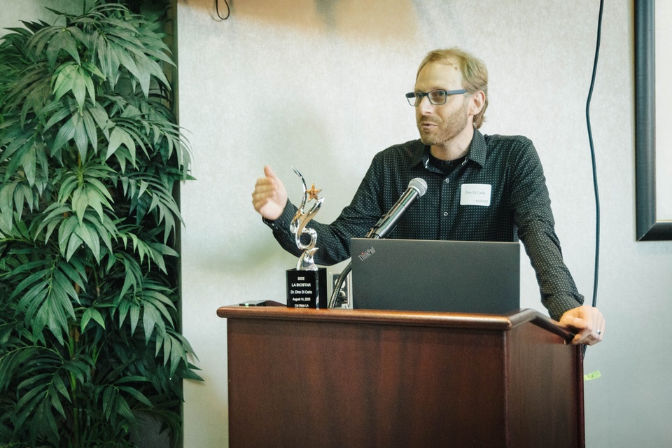 A speaker stands at a podium giving remarks during a Cal State LA Biostart event, with an award trophy and laptop on the podium and a potted plant in the background.