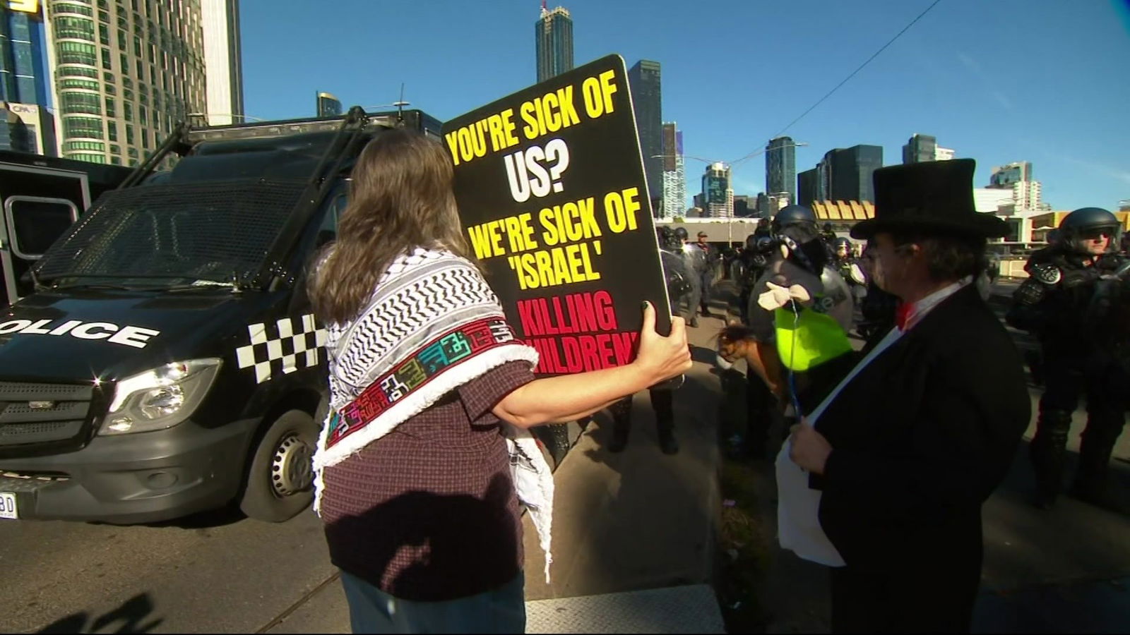 A woman with a black and white scarf on her shoulders holds a sign up at police officers.