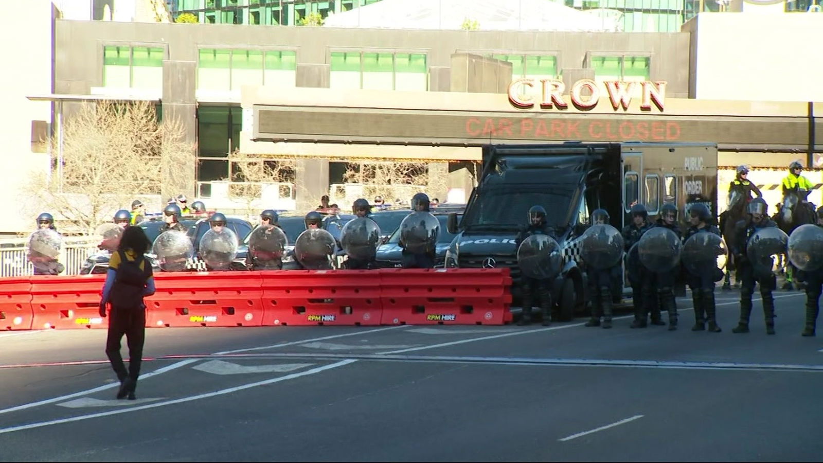 Police dressed in black with helmets and shields stand behind an orange barricade beside a black police van.