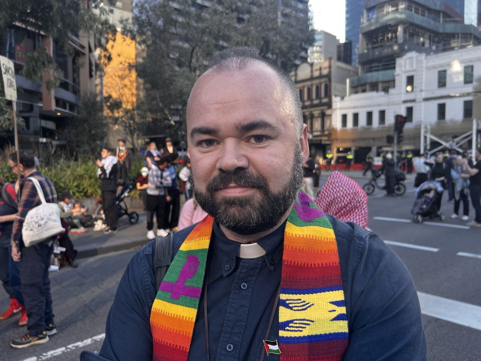 A man in a black button-up shirt with a religious collar, wearing a multi-coloured scarf