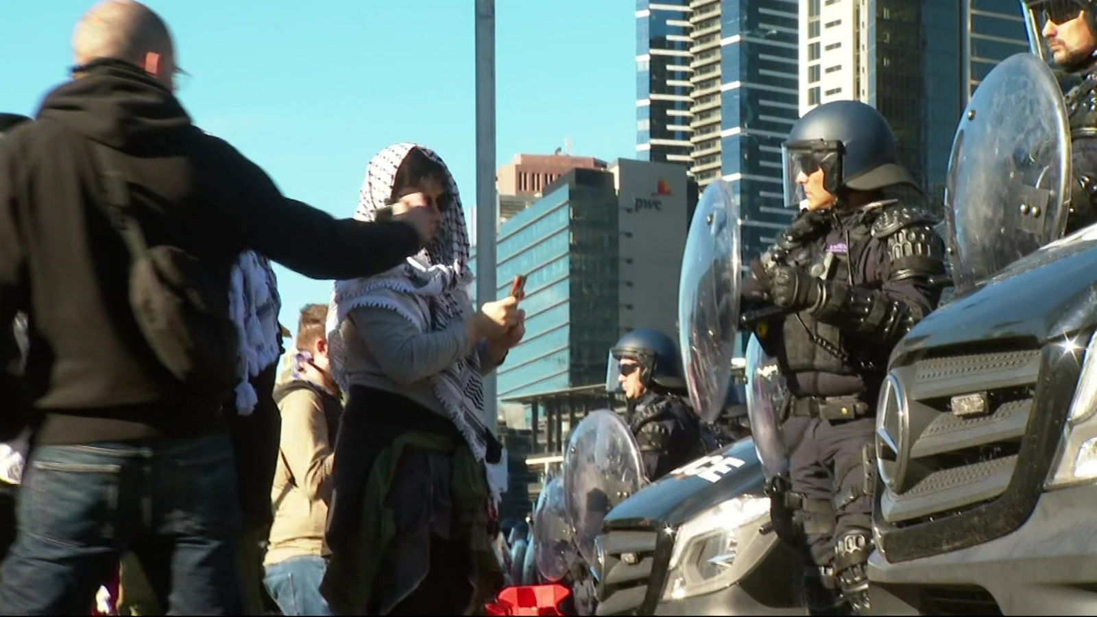 A man and a woman in a black and white scarf hold phones up taking pictures of riot police.