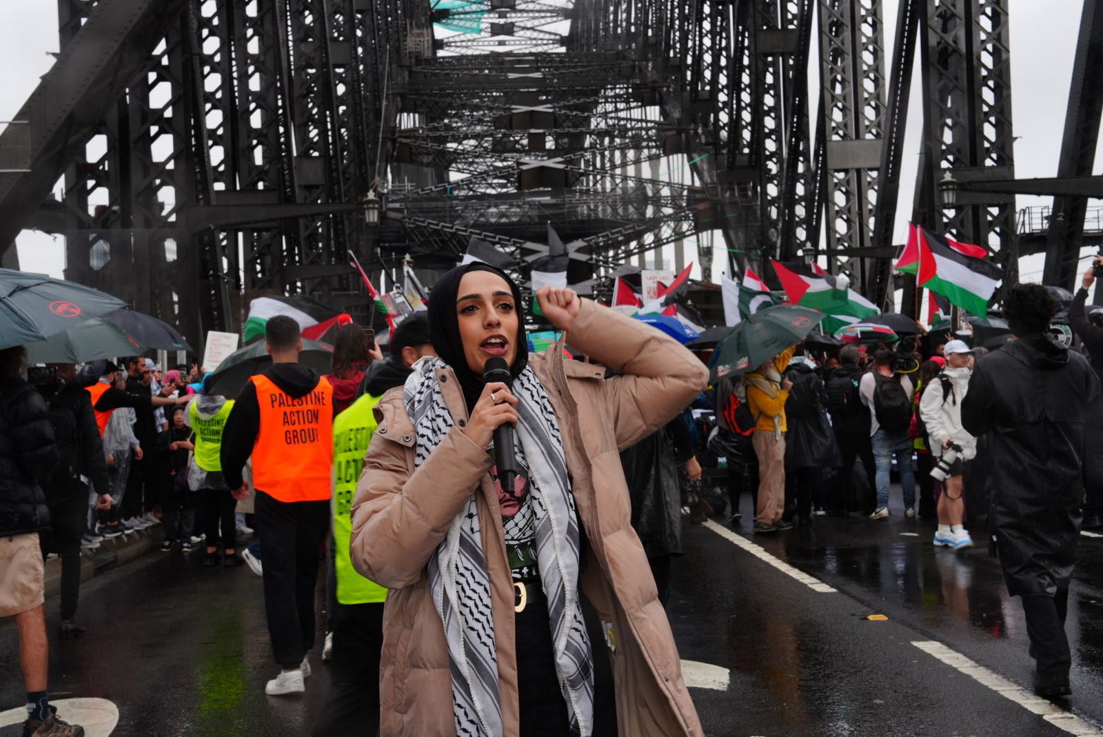 A woman wearing a beige puffer and keffiyeh speaks into a microphone with a raised fist
