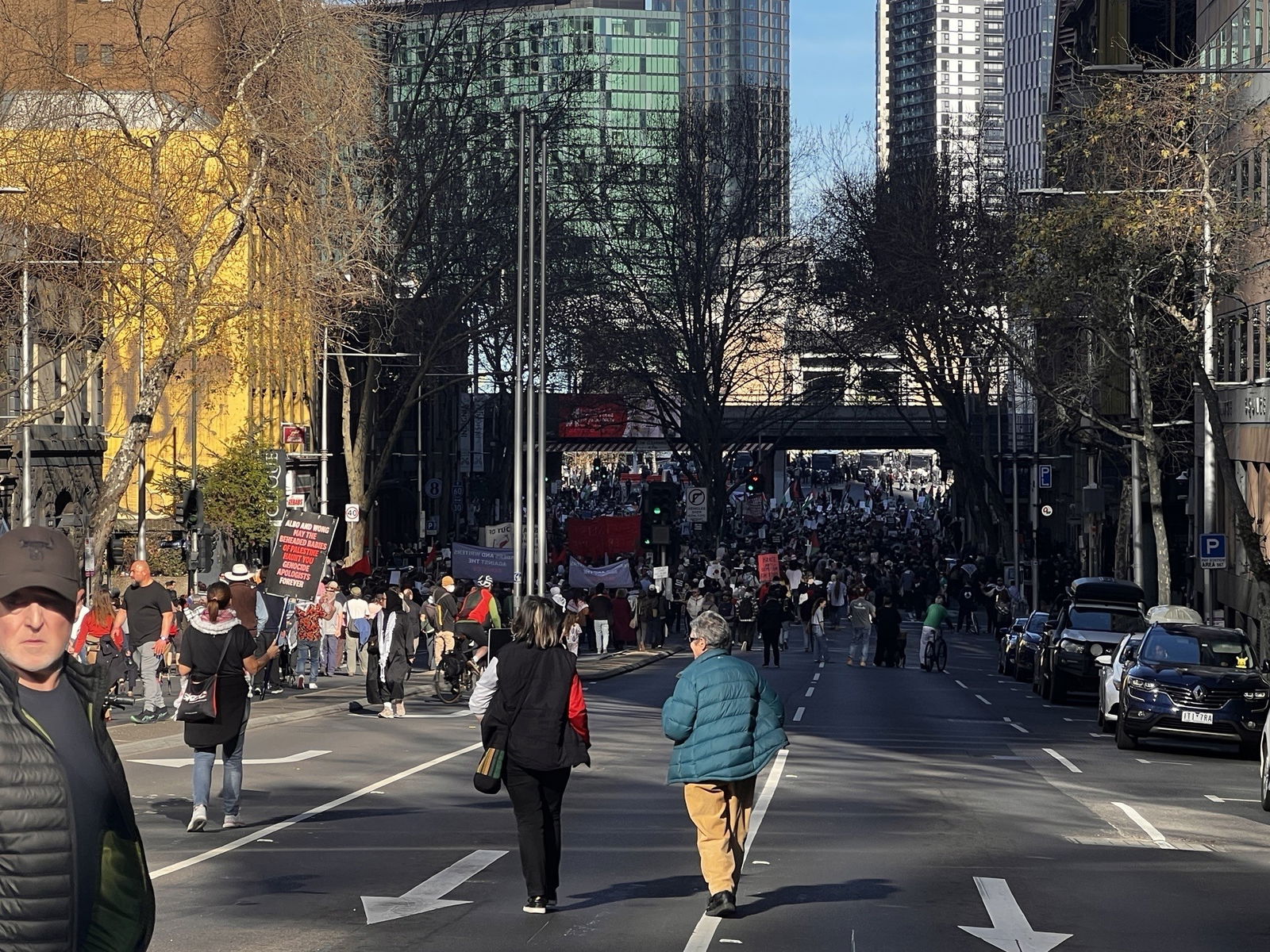 A large crowd moves down the middle of a CBD street and under a bridge in the distance.