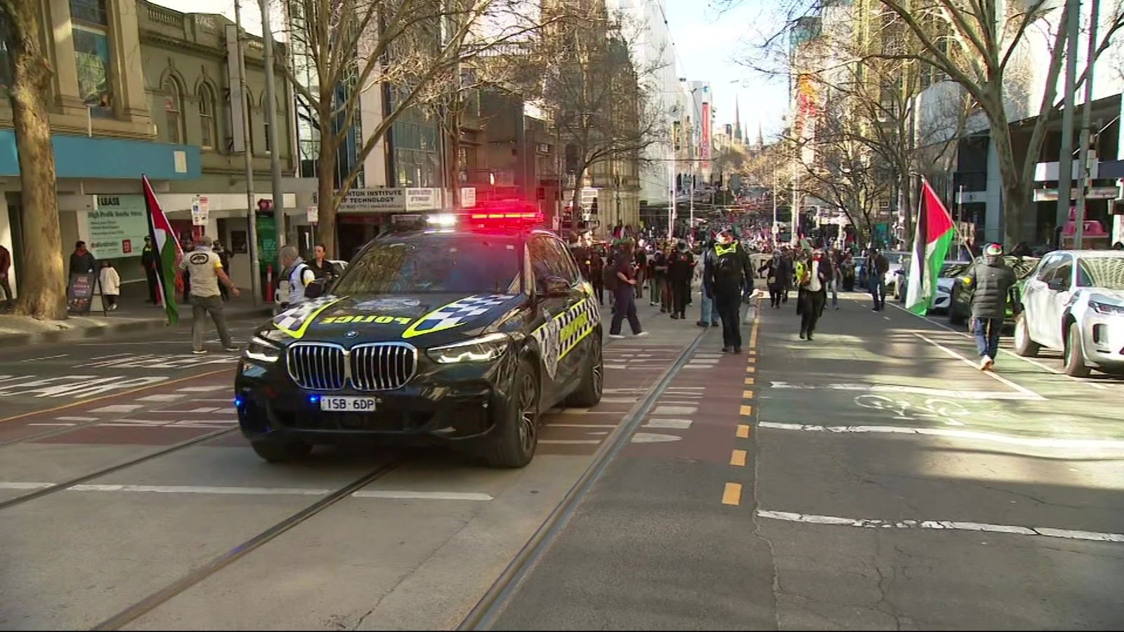A police car with its lights on drives in front of a large crowd walking down a city street.