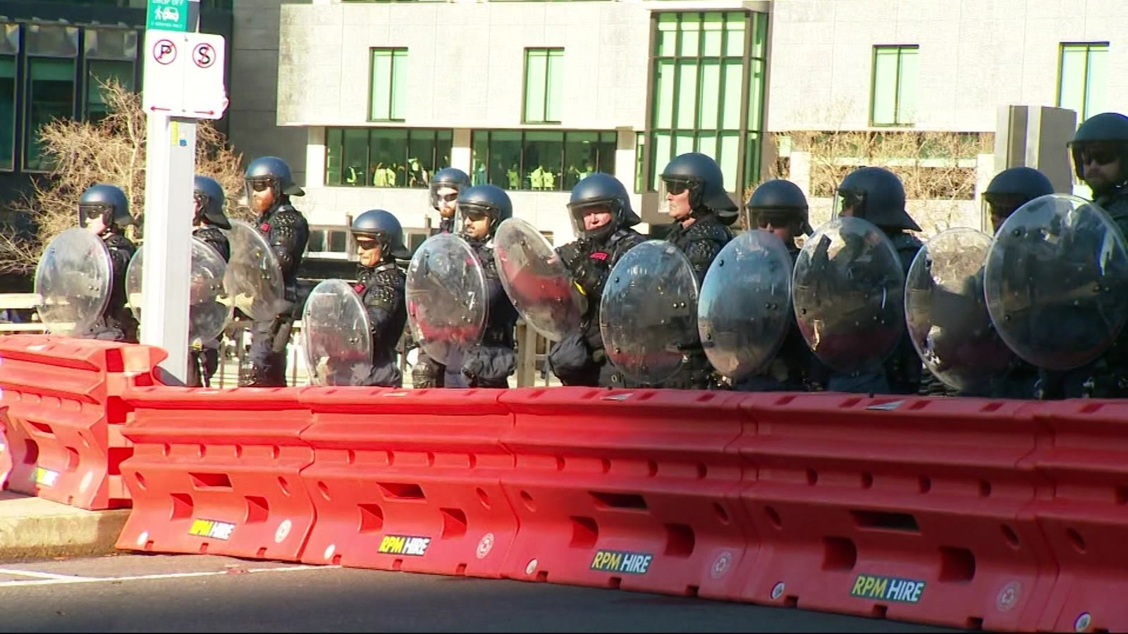 Police in helmets holding shields stand behind an orange barricade.