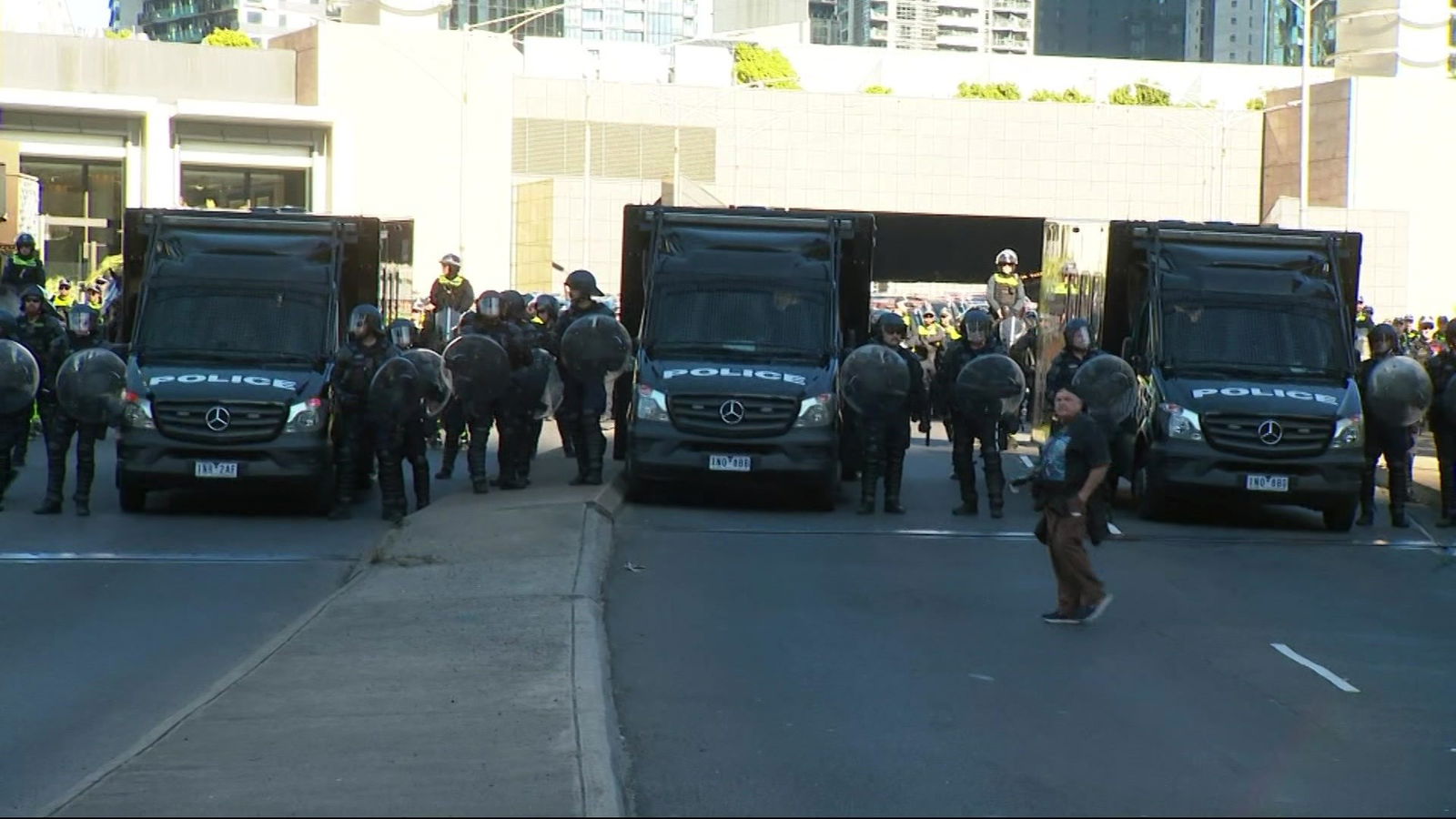 Police in helmets holding shields stand between three black vans on a street.