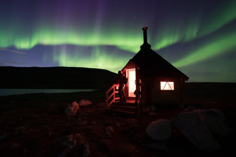 A person stands in the doorway of a small, warmly lit cabin under a night sky illuminated by vivid green Northern Lights, with a hill silhouetted in the background.