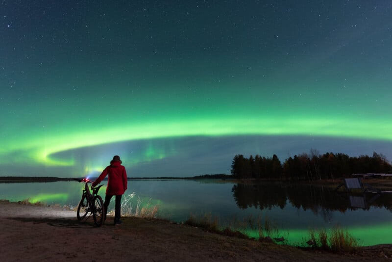 A person in a red jacket stands beside a bicycle on a lakeshore at night, gazing at a vibrant green aurora borealis illuminating the sky and reflecting on the calm water.