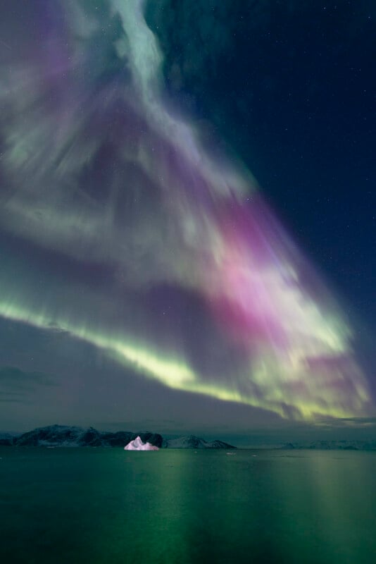 Colorful aurora borealis with green and purple lights spreads across a starry night sky above calm water and a distant, snow-covered mountain range. An iceberg glows in the lower center.