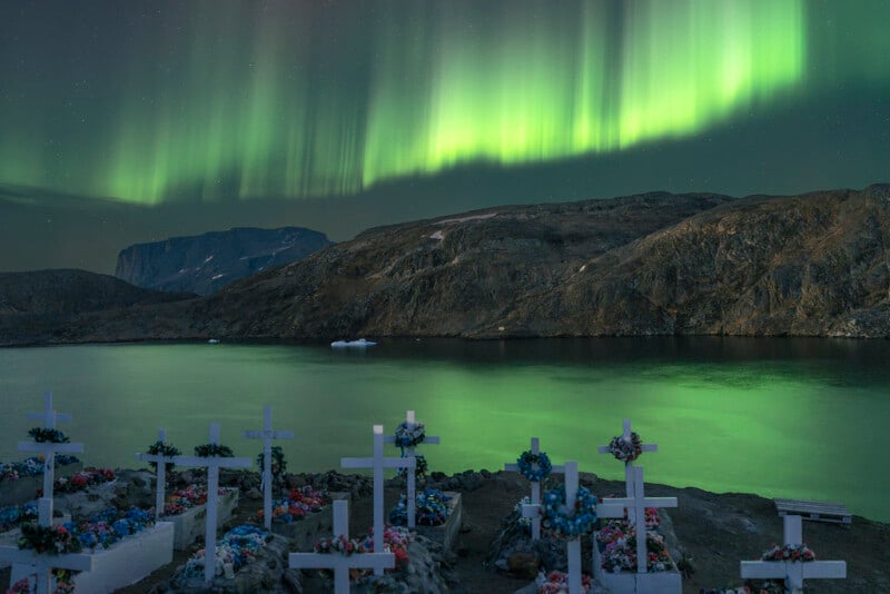 Rows of white crosses in a cemetery overlook a calm body of water and rocky hills, with green aurora borealis lights illuminating the night sky above.