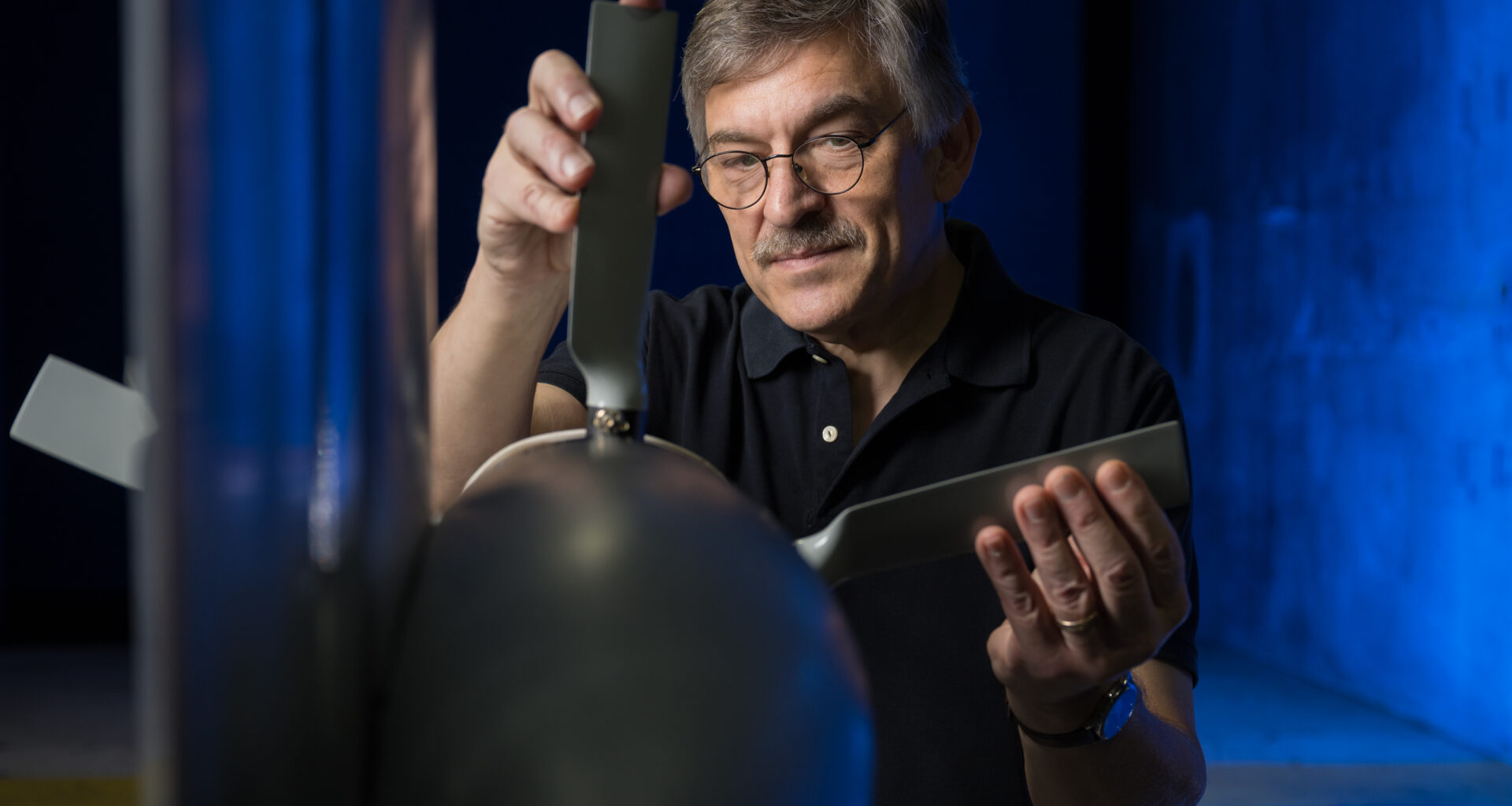 A man is adjusting a propeller blade attached to a test model, with a blue-lit background.