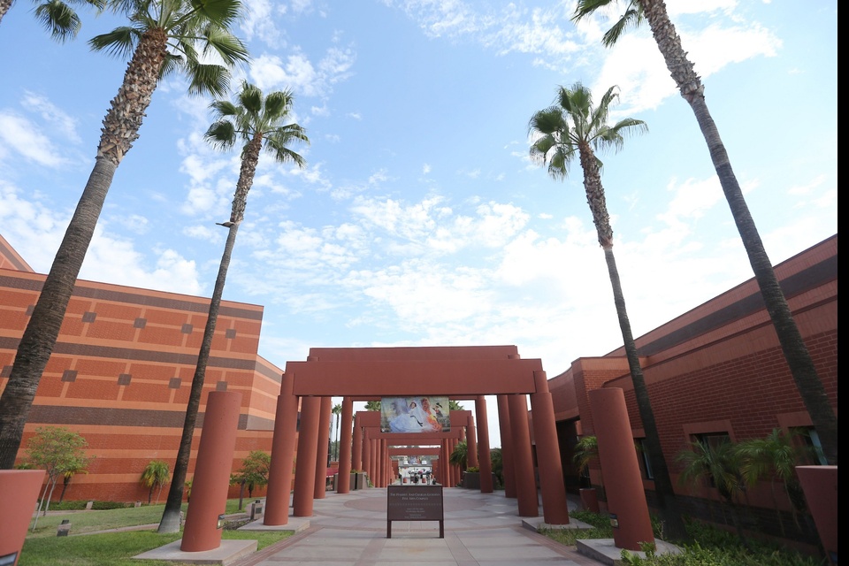 Entrance to the Luckman Fine Arts Complex at Cal State LA, featuring symmetrical red pillars, palm trees, and bright blue skies with scattered clouds.