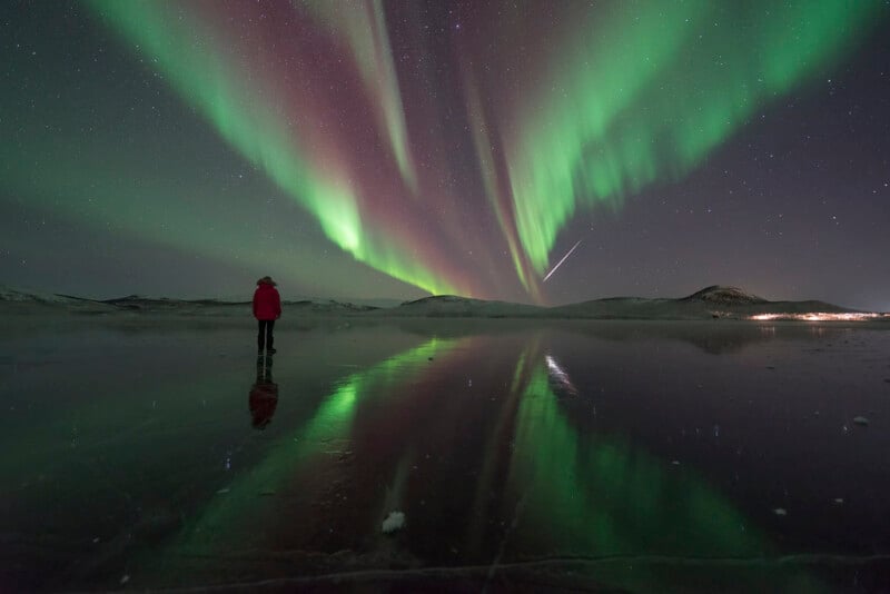 A person in a red jacket stands on a reflective frozen lake, watching vibrant green and purple northern lights dance across the night sky, with a shooting star visible.