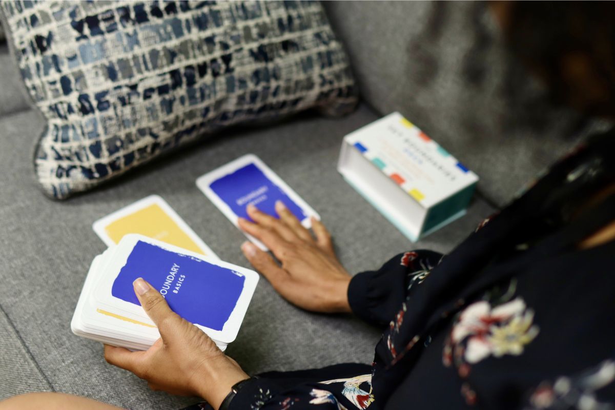 A person sits on a gray sofa, holding and sorting through a deck of colorful cards, with a box for the cards placed nearby.