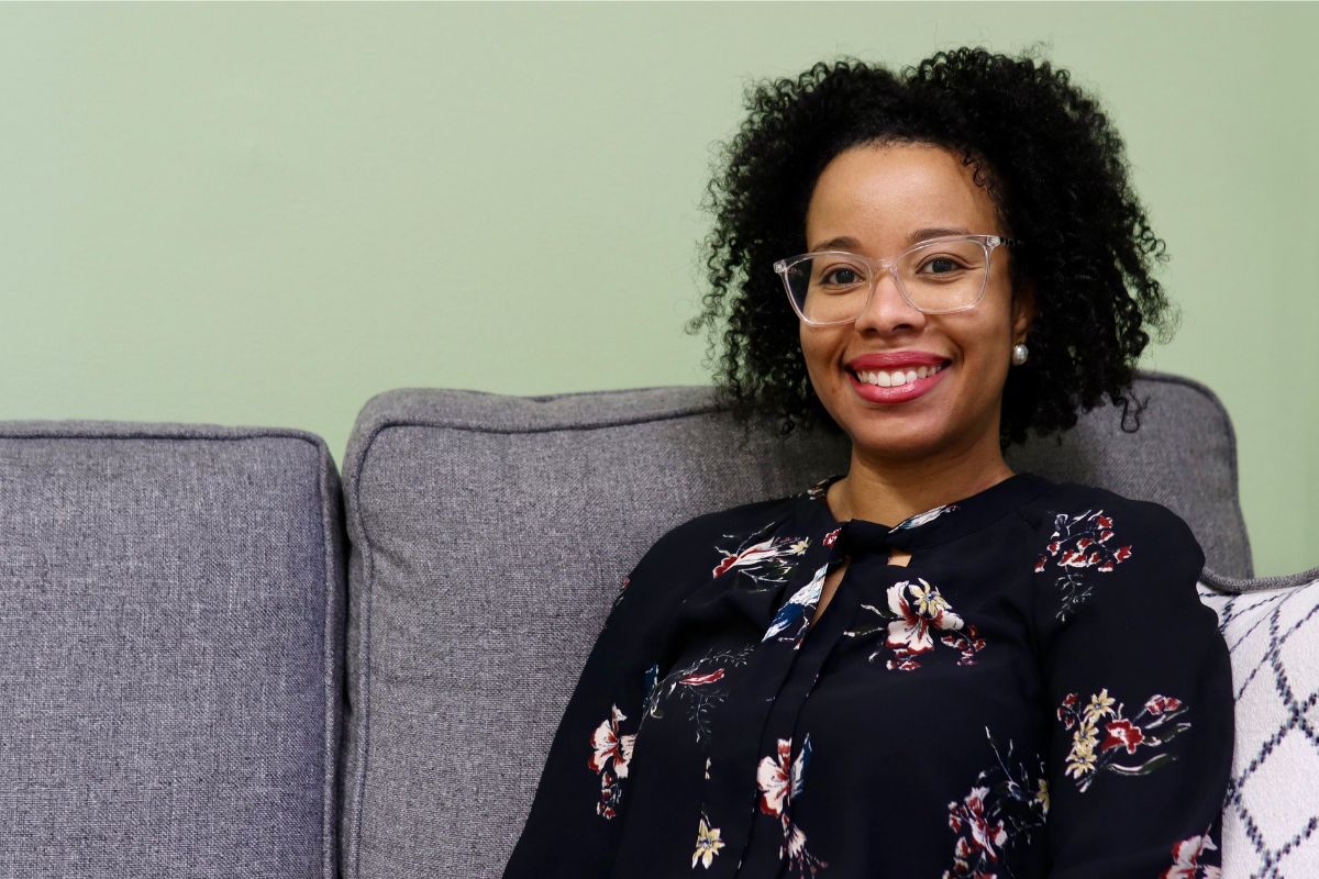 A woman with curly hair and glasses sits on a gray couch, smiling, wearing a dark floral blouse against a light green wall.