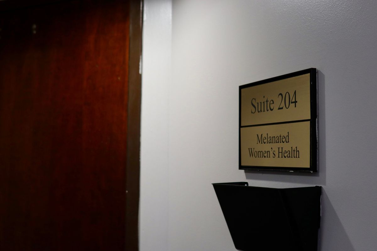 A wall-mounted sign reads "Suite 204 Melanated Women’s Health" next to a dark door and an empty black file holder.