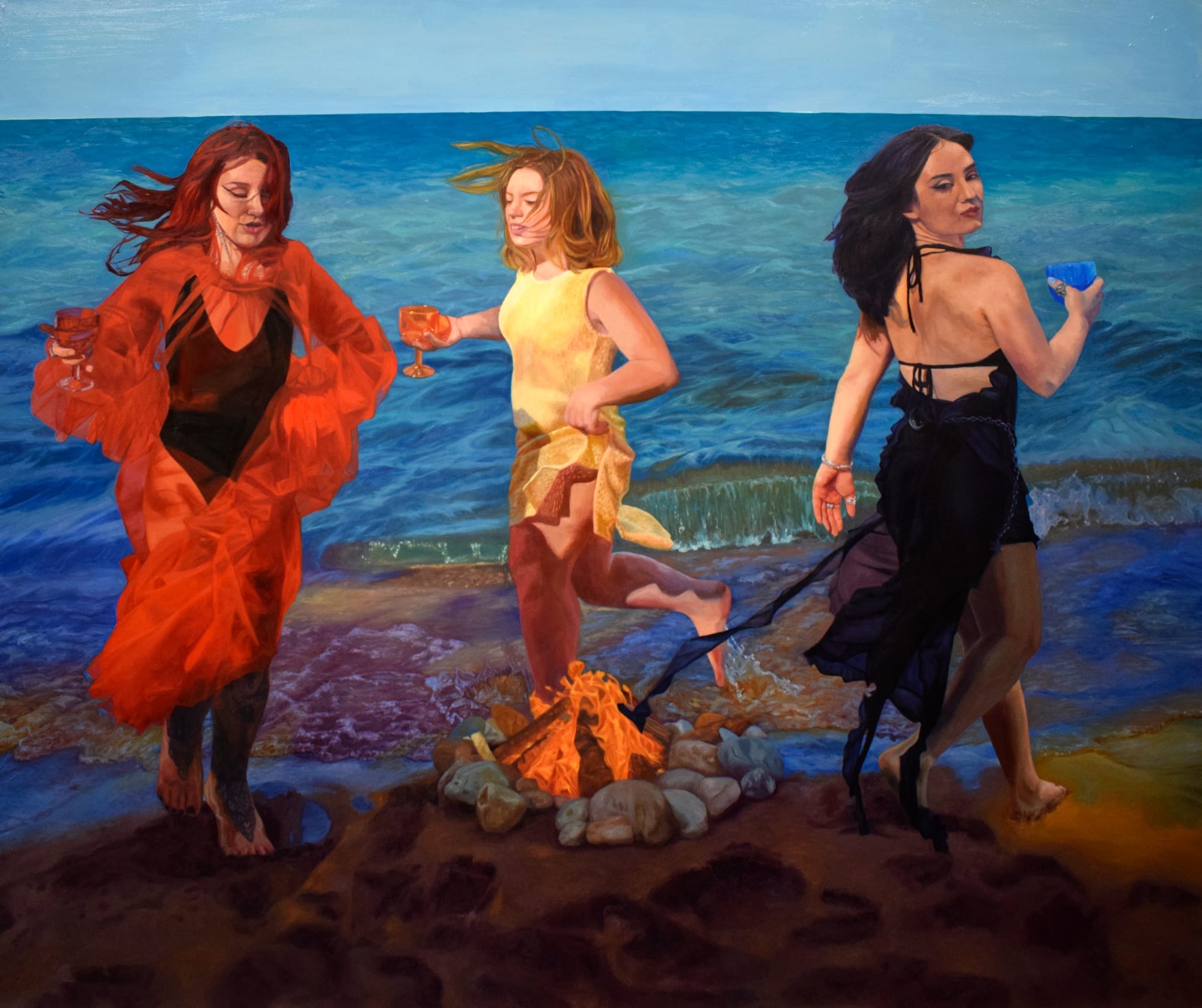 three women in red, white, and black dance around a fire on a beach