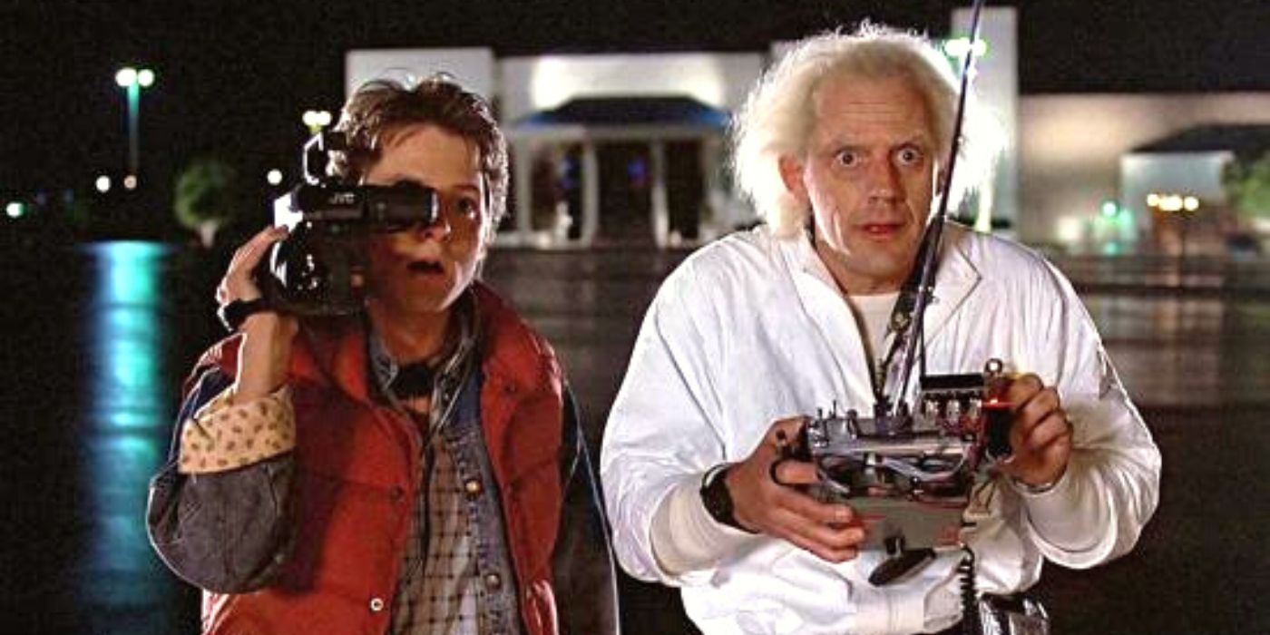 Marty (Michael J Fox) holds a video camera & stands next to Doc (Christopher Lloyd), who holds a remote control, in Back to the Future (1985).