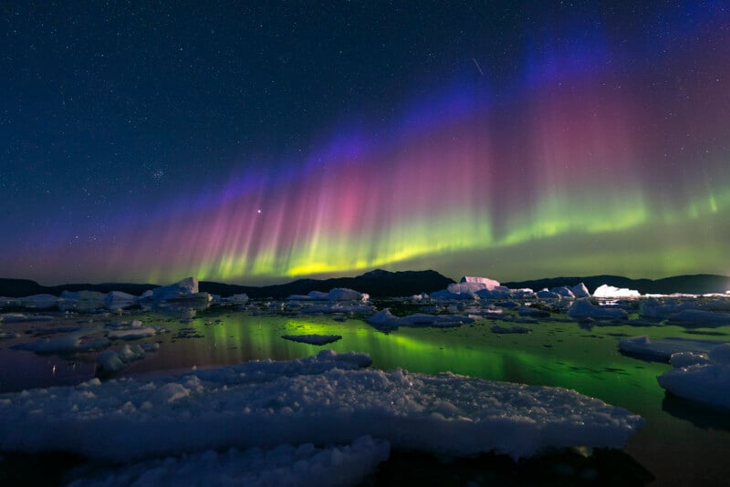 Colorful aurora borealis lights in purple, pink, and green illuminate the night sky above icy water with floating icebergs and snow, reflecting the vibrant lights. Mountains are visible on the horizon.