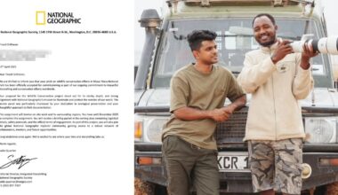 Two men stand smiling and talking in front of a safari jeep. On the left, one man wears a tan t-shirt and green pants; on the right, the other man wears a beige shirt and camouflage shorts. A National Geographic letter is visible on the left side.