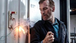 A man with cuts on his face and wearing a worn jacket holds a lighter to a makeshift explosive device on a wall. Sparks fly as the wires ignite, casting light on his tense, focused expression. The setting appears industrial, gritty, and dangerous, hinting at a high-stakes action scenario.