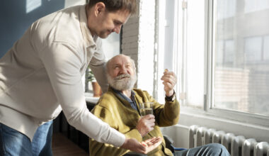 A young man giving medicine to an older man in a wheelchair. Caregiving featured image