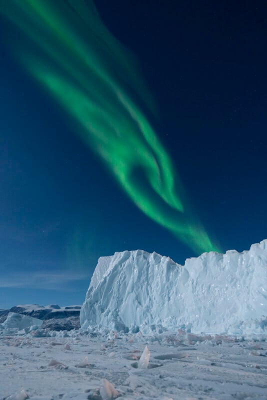 Green Northern Lights illuminate a clear night sky above a large, jagged iceberg in a snowy, icy landscape.