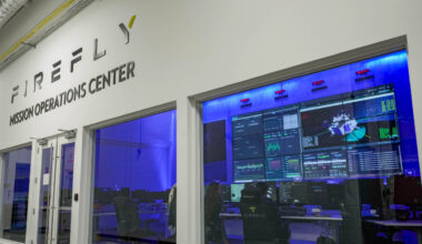 A control room with blue lights and lots of computer panels sits behind glass with a sign that reads Firefly Mission Operations Center above it.