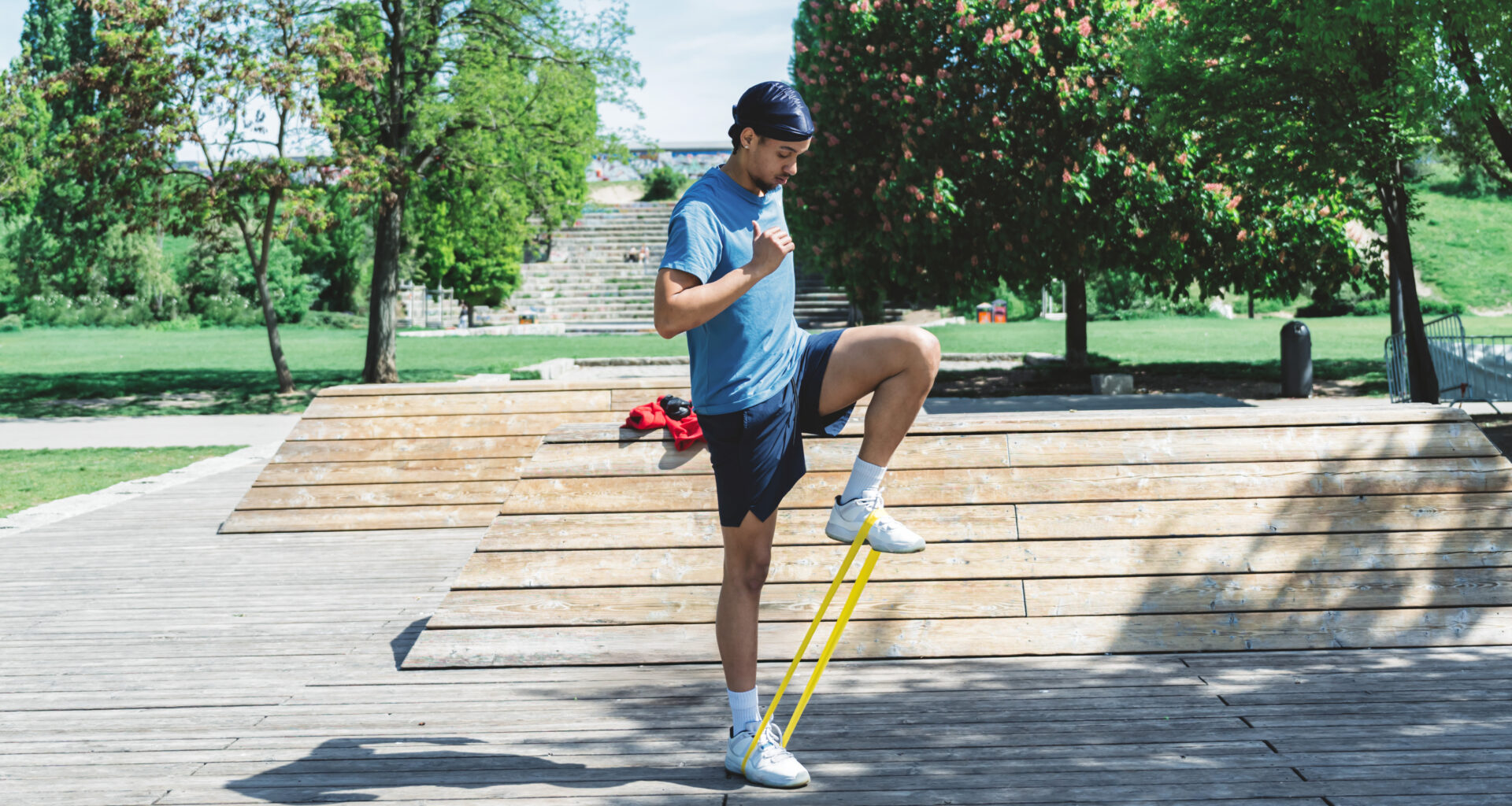 A man performs a resistance band exercise in a park outdoors. He is standing, with the band looped around his feet, and one knee bent so that he is in a marching pose. Behind him we see a long path, steps and trees.