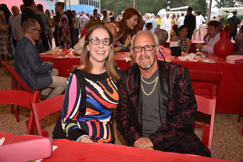 Paul herman in a black jacket and grey shirt with two necklaces and dark glasses sitting next to a woman in a colorful dress at an event