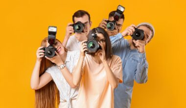 Five young adults stand close together against a bright yellow background, each holding a camera up to their face as if taking a photo. They appear cheerful and engaged in photography.