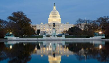 The U.S. Capitol building illuminated at dusk is reflected in a calm pool of water, surrounded by trees and statues in Washington, D.C.