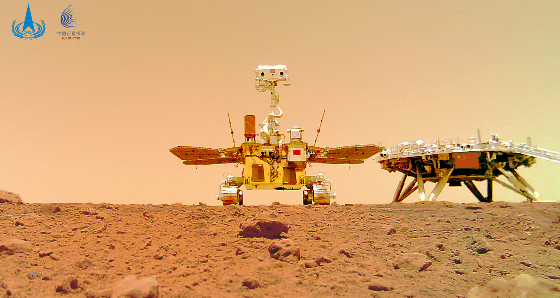 A photo of a metal rover with wheels and two solar panels standing on reddish brown dirt.