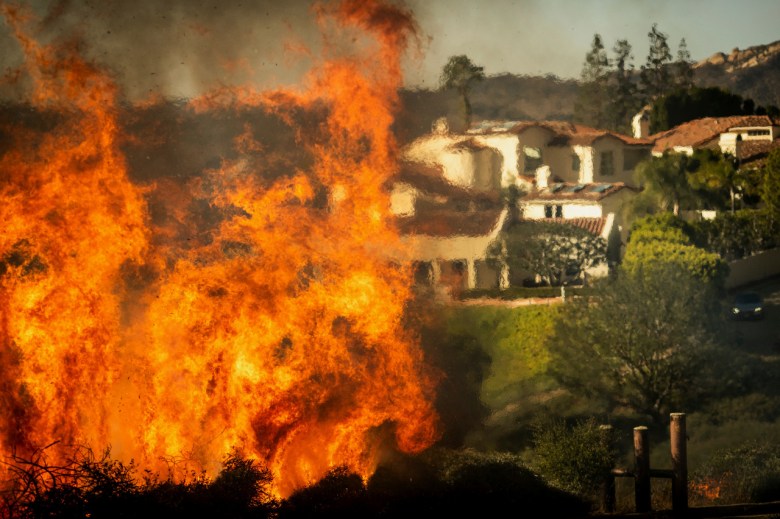 A wildfire with tall orange flames near homes with white walls and red-tiled roofs.