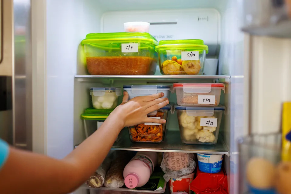 A person organizes a fridge with labeled containers holding various foods, demonstrating meal prep and organization for efficient work-week planning