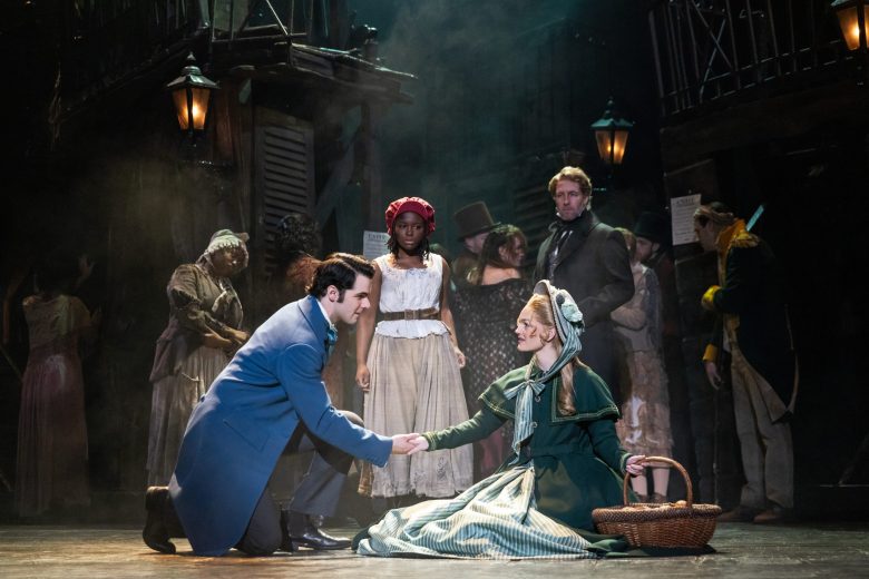 A student offers a hand to a fallen actress in historic French dress with a crowd of dirty actors behind them. 