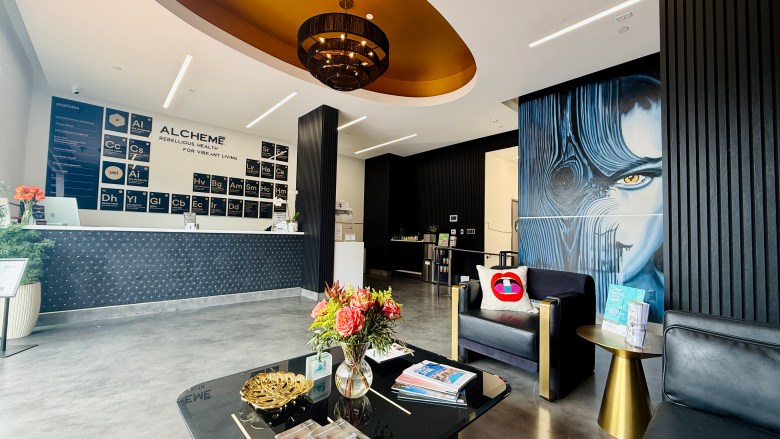 A room with modern furnishings, a mural of a person on the wall to the right and the word "Alcheme" shown on the far wall behind a reception desk. 