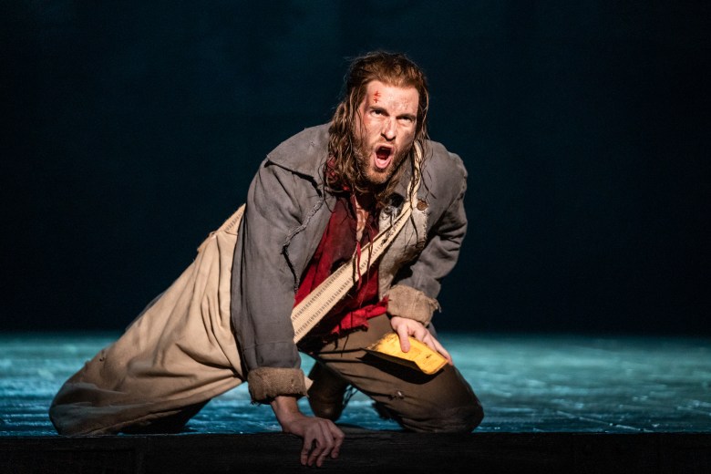 An actor in ragged clothes with a cut on their forehead kneeling while singing as part of the “Les Misérables” production.