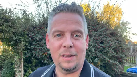 STEVE BEECH/BBC NEWS Man with goatee beard in dark shirt in front of a hedge