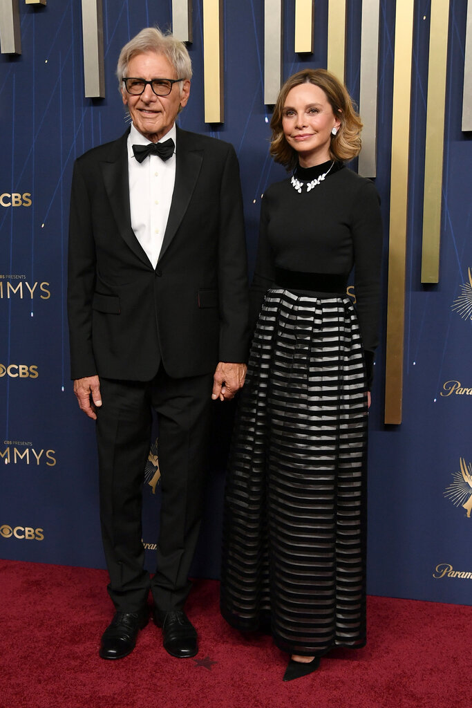 A man wearing a black suit (left) holds hands with a woman wearing a black dress with a striped skirt.