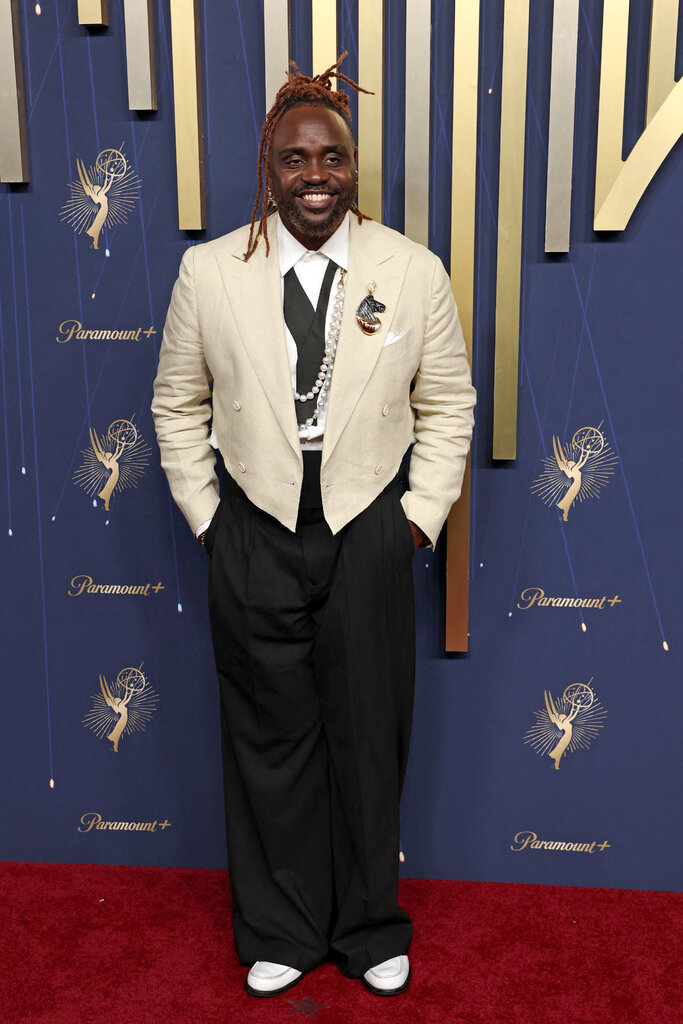 A man wears a tuxedo with a cream jacket and black pants and several strands of pearls clipped to his collar.