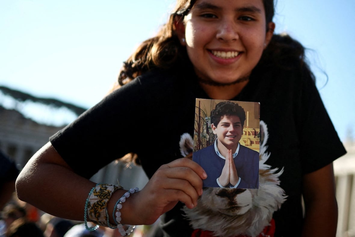 A smiling teenage girl holds up a photo of a curly-haired teenage boy smiling and making prayer hands