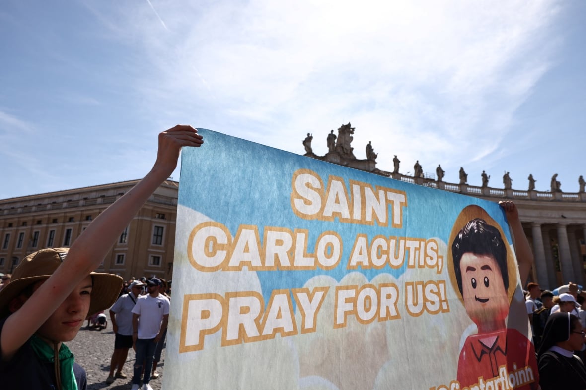 People hold up a banner with a Lego man with curly brown hair and the words "Saint Carlo Acutis, pray for us!" in big bubble letters. 